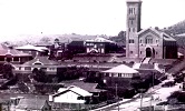 Historic black‑and‑white view of a town with a prominent church building, surrounding houses, and streets in the foreground