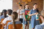 Students seated in a church while 3 students walk with candle, book and cross.