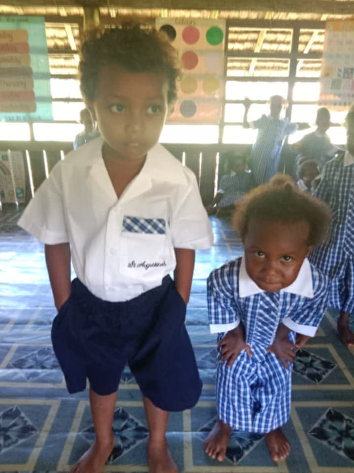 Two young students in school uniforms standing at the front of a classroom with other students in the background