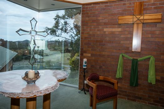 Small chapel space with wooden cross on brick wall, altar table, and window with decorative glass cross.